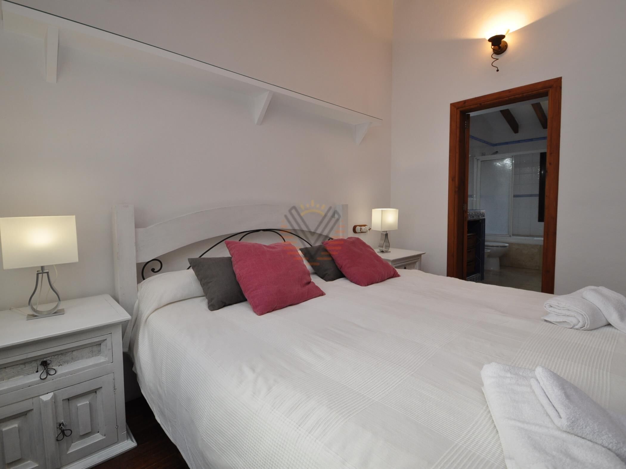 Ein ordentlich bezogenes Bett mit weißen Leinen und zwei Kissen, ein Nachttisch mit Lampe, ein kleiner Schrank und eine Wandleuchte.