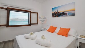 A bedroom with a bed, a nightstand, a window with a view of the sea, and a painting of a lighthouse. - Sa Marina 7A, villa direct on the beach (Alcudia - Puerto de Alcudia)