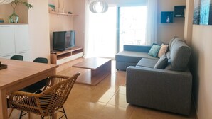 Una sala de estar moderna con un sofá gris, una mesa de centro de madera, un televisor sobre un mueble, una silla de mimbre y una mesa de comedor con sillas.