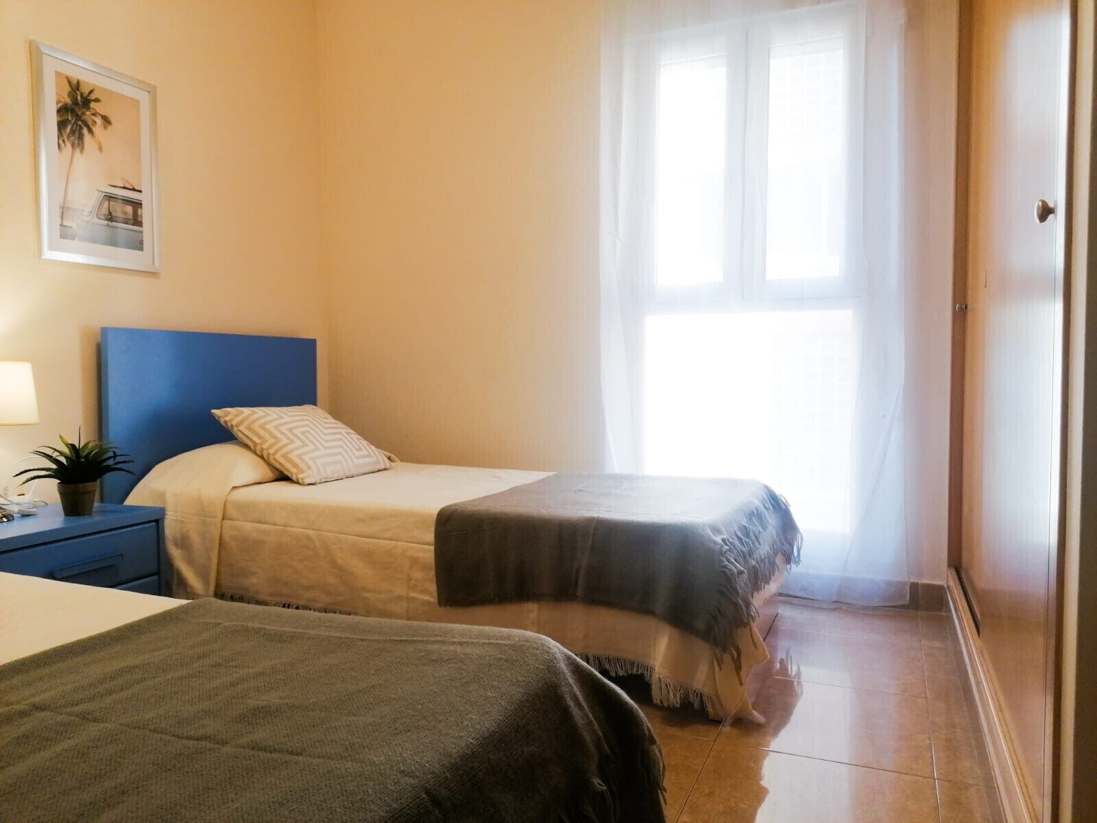 Hotellihuone, jossa on kaksi sänkyä, yöpöytä, lamppu ja ikkuna, jossa on läpikuultavat verhot.