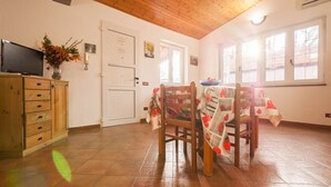 Una habitación con techo de madera, piso de baldosas, una mesa de comedor con sillas, un televisor sobre un mueble y una puerta con un letrero.