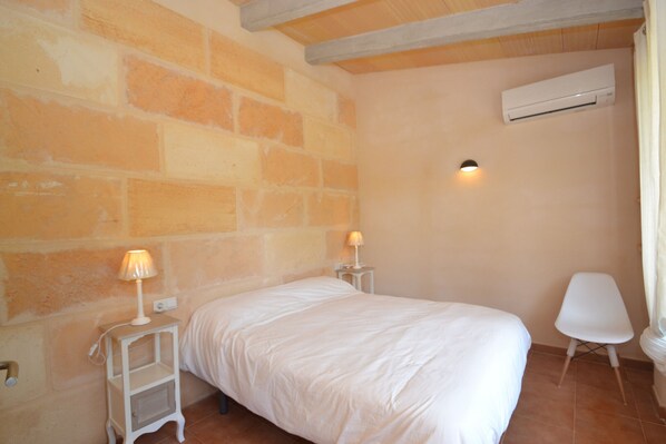 A bedroom with a bed, two bedside tables with lamps, a white chair, and an air conditioning unit on the wall.