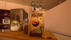 Una botella de café de Tarrazú con dos dosis, sobre una tabla de madera junto a un tarro de vidrio con azúcar y un pequeño recipiente con granos de café.