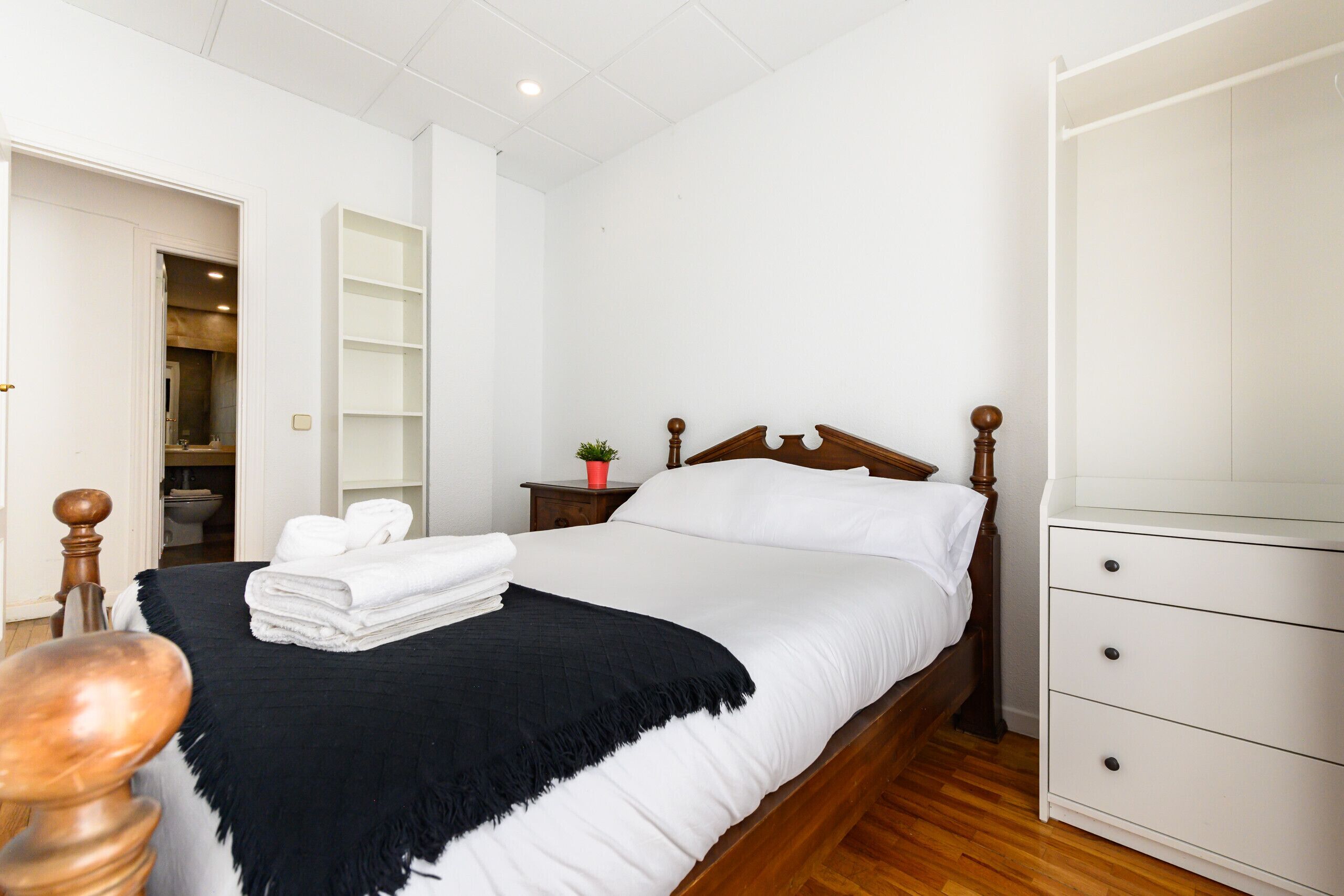 Un dormitorio con cama, mesita de noche, cómoda, armario y baño visible a través de una puerta abierta.