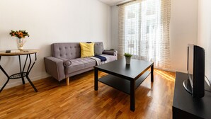 Ein modernes Wohnzimmer mit grauem Sofa, einem kleinen Tisch, einem Flachbildfernseher und einem großen Fenster mit leichten Vorhängen.