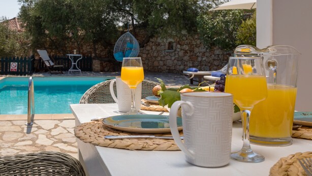 Un coin petit-déjeuner aménagé au bord de la piscine, avec une table, des assiettes, des verres et une carafe de jus d’orange.