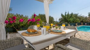 Une table à manger installée à l’extérieur, avec un assortiment de produits pour le petit-déjeuner, une piscine en arrière-plan et une végétation luxuriante.