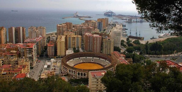 A coastal cityscape with a prominent bullring in the foreground, surrounded by high-rise buildings and a port with ships and cranes. - Victoria Premium Apartment (Málaga)
