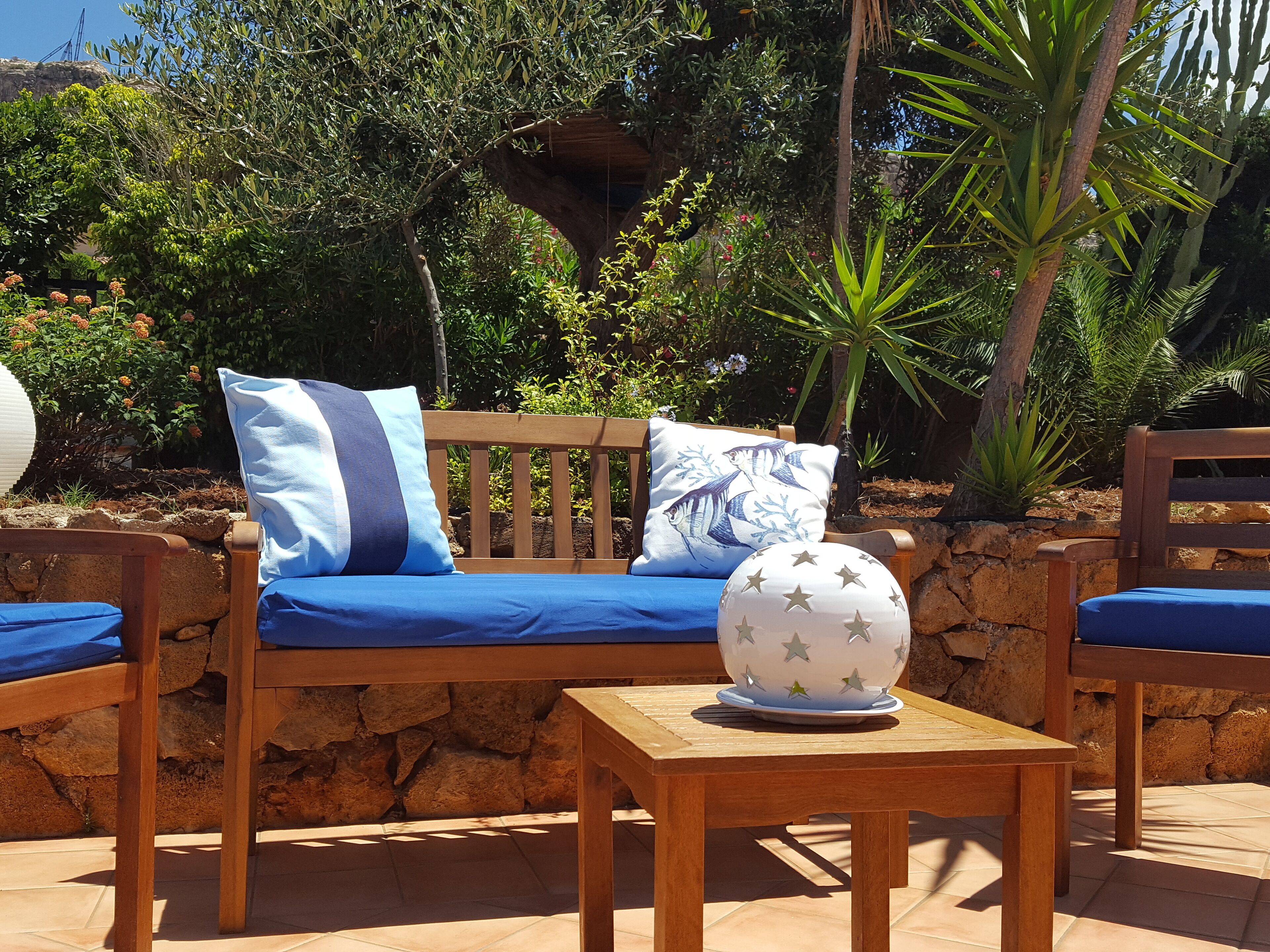 A wooden bench with blue cushions and pillows, a round table with a star-patterned bowl, and a stone wall backdrop with various plants.