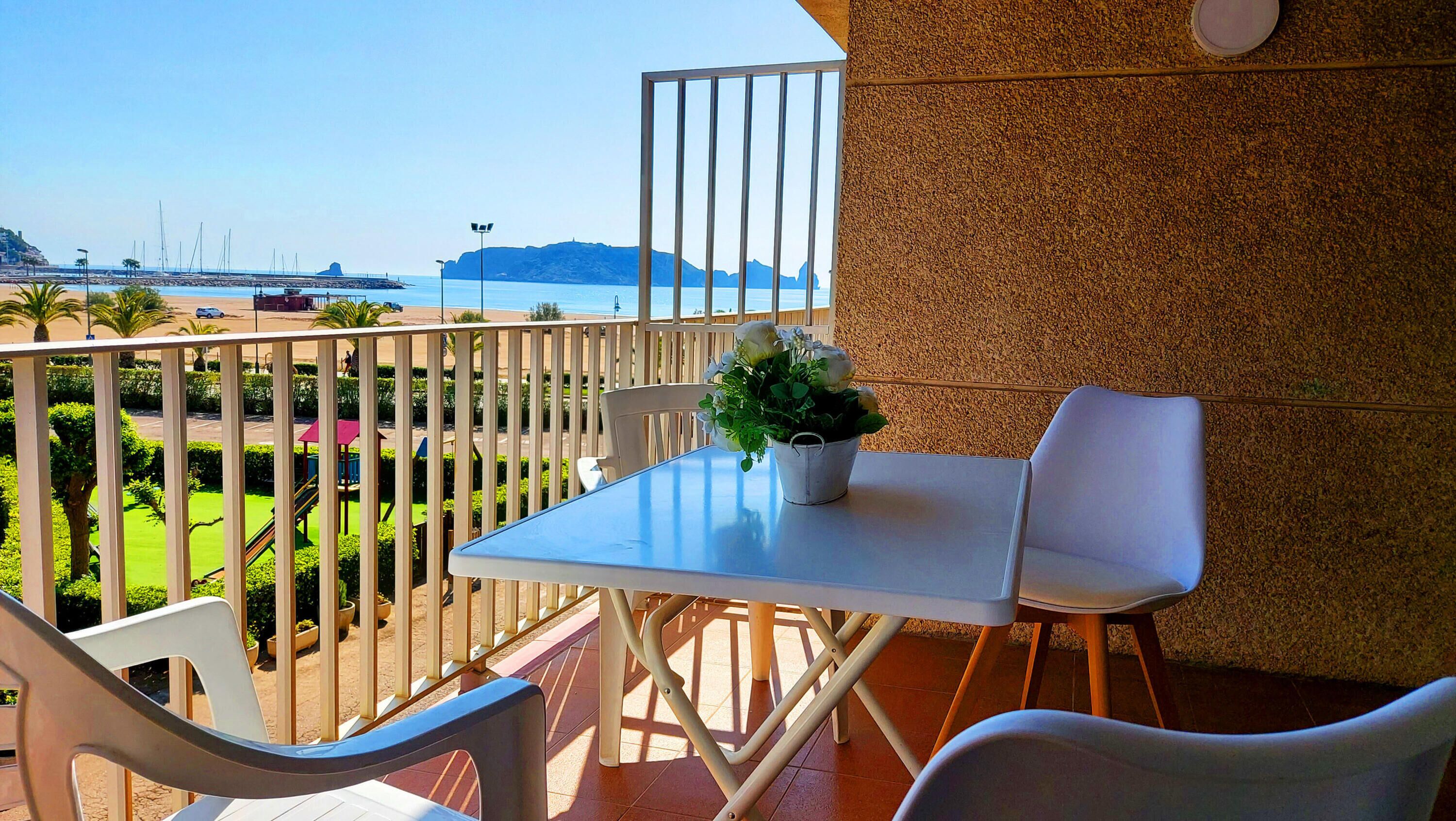 Un balcón con mesa y sillas, una planta en maceta y vistas al mar y a la costa lejana.