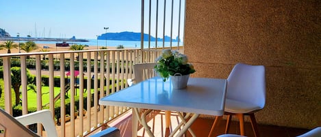 A balcony with a table and chairs, a potted plant, and a view of the sea and distant land.