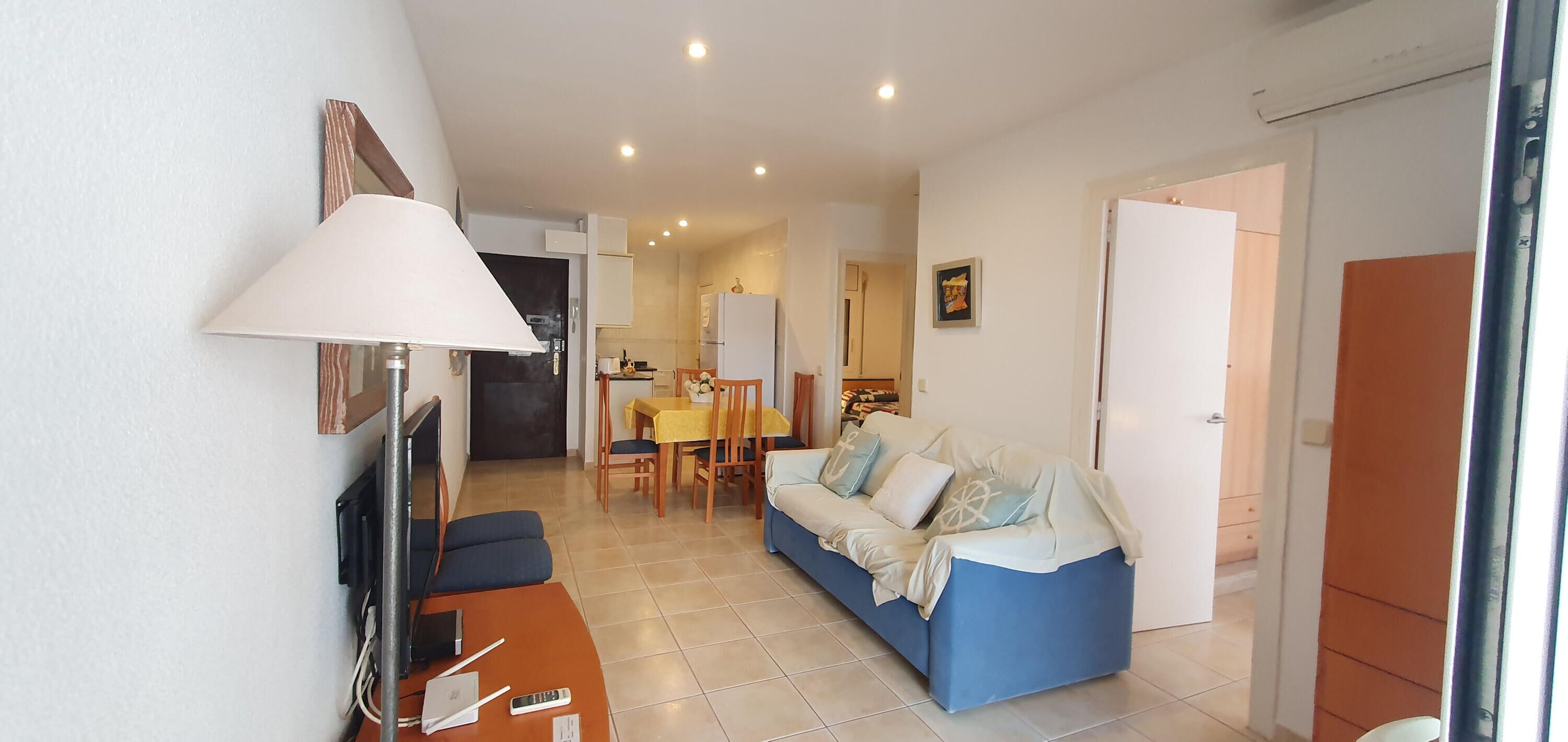 Una sala de estar con sofá, televisión, lámpara, mesa de comedor con sillas y una cocina al fondo.