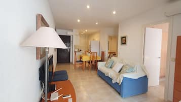 Una sala de estar con sofá, televisión, lámpara, mesa de comedor con sillas y una cocina al fondo.
