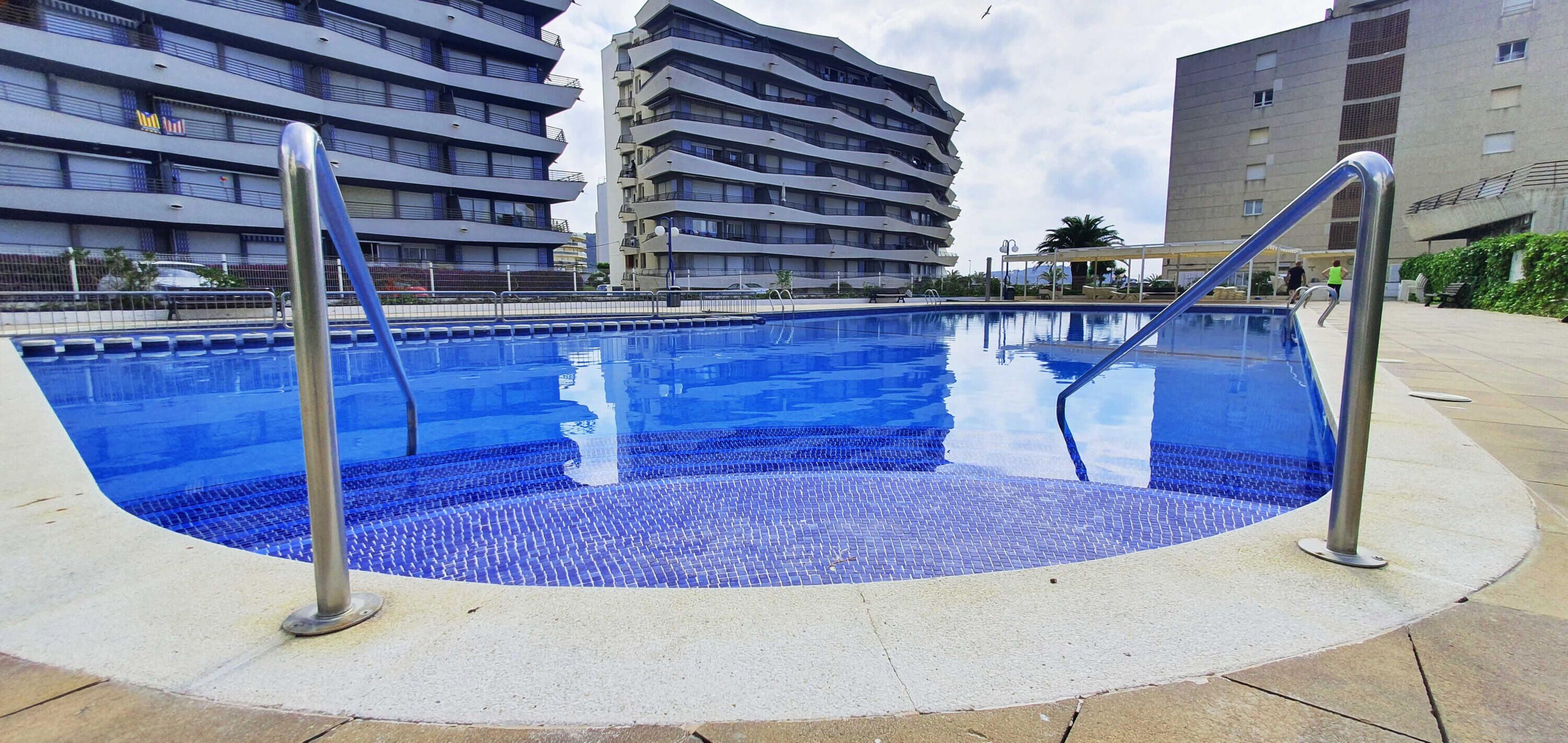 Una piscina con borde de baldosas, rodeada por una solera de concreto, y contigua a edificios residenciales de varios pisos.