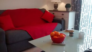 A modern living room with a grey sofa, red cushions, a glass coffee table with a bowl of oranges, and a decorative vase. - T1 standing entrance of CALVI  (Calvi)