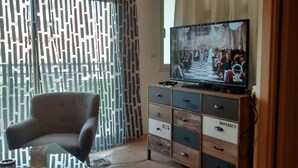 A living room with a television on a wooden cabinet, a grey armchair, and patterned curtains. - T1 standing entrance of CALVI  (Calvi)