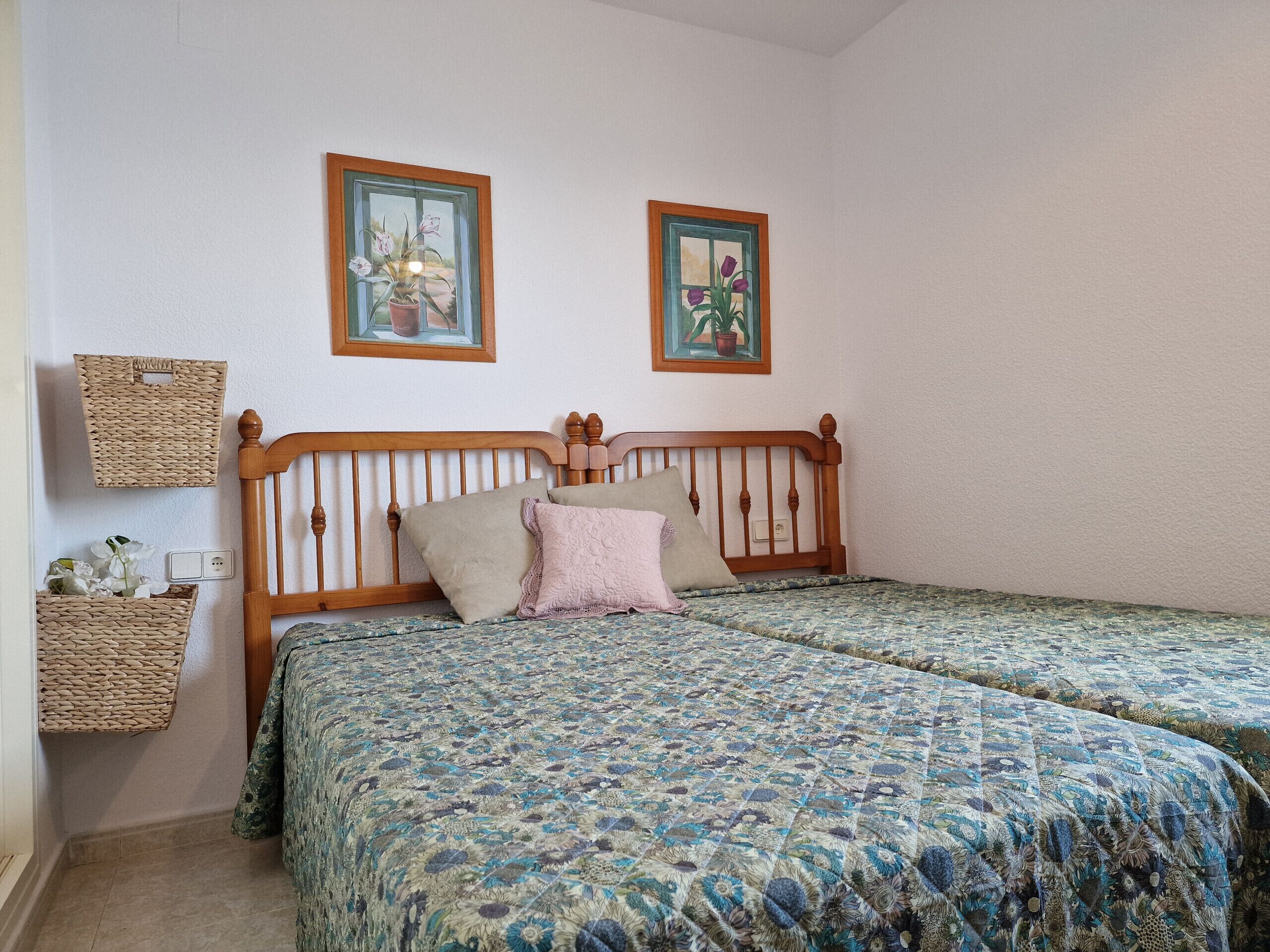 A bedroom with two beds, a wooden headboard, a floral bedspread, and two framed pictures on the wall.