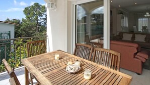 Une table en bois avec des chaises sur un balcon, une porte vitrée donnant sur un espace intérieur, et une vue sur des arbres et un bâtiment au loin.