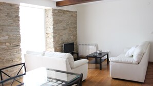 Een woonkamer met een stenen muur, witte banken, een glazen tafel en een televisie.