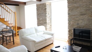 Een woonkamer met een stenen muur, een witte bank, een tv op een standaard, een houten tafel en een trap.