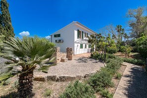 A white building with a sloped roof, air conditioning units, and a garden with various plants and a palm tree.