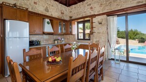 A stone-walled kitchen with wooden cabinets, a dining table with chairs, a refrigerator, and a view of a swimming pool.