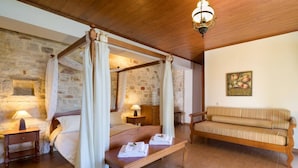 A four-poster bed with white linens, a wooden bench with towels, a wooden sofa, and a stone wall.