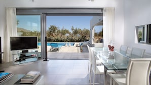 Un salon moderne avec une table en verre, des chaises blanches, un téléviseur à écran plat et une vue sur une piscine et des arbres.