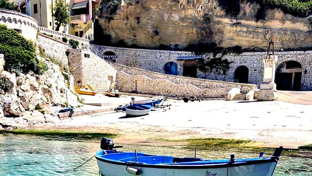 Una barca ancorata in acque limpide e poco profonde, con una riva rocciosa e edifici sullo sfondo.