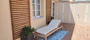 A sunlit outdoor seating area with a cushioned bench, wooden slatted walls, and a potted plant.