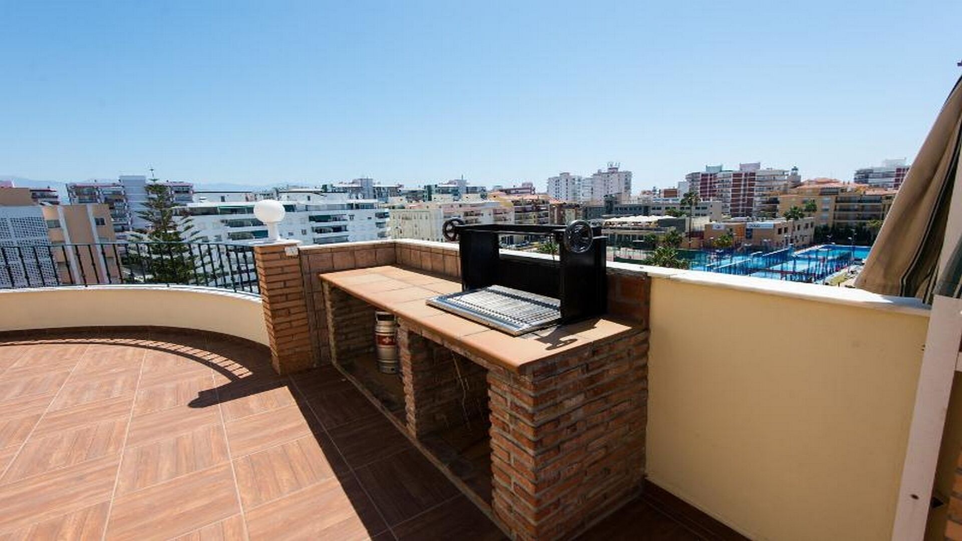Un balcon avec un barbecue et une table, donnant sur un paysage urbain avec des immeubles et une piscine.