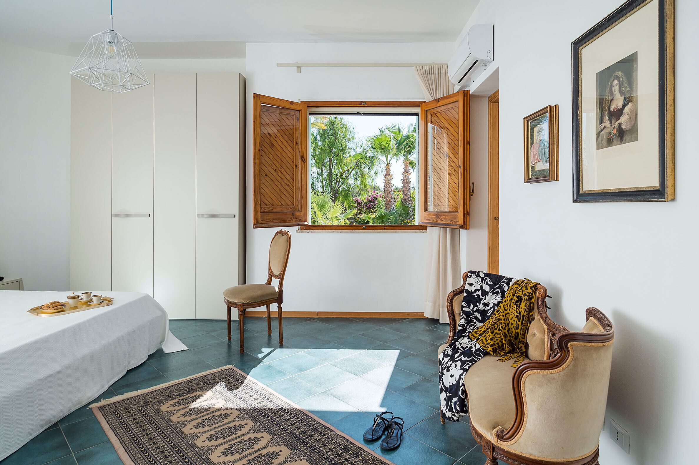 A bedroom with a bed, a chair, a sofa, a window with open shutters, and a view of greenery outside.