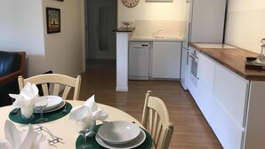 A dining area with a table set for two, a kitchen with white appliances, and a clock on the wall.