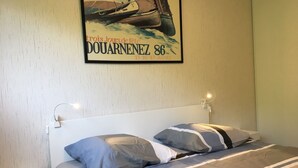 A neatly made bed with striped bedding and two pillows, a wall-mounted light, and a framed poster of a sailboat.