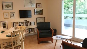 A living room with a dining table, chairs, a bookshelf, a leather armchair, a TV, and a large window with a view of greenery.