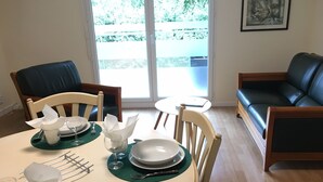 A dining area with a table set for two, chairs, a glass of water, a plate, and a napkin. There is a window with a view of greenery outside.