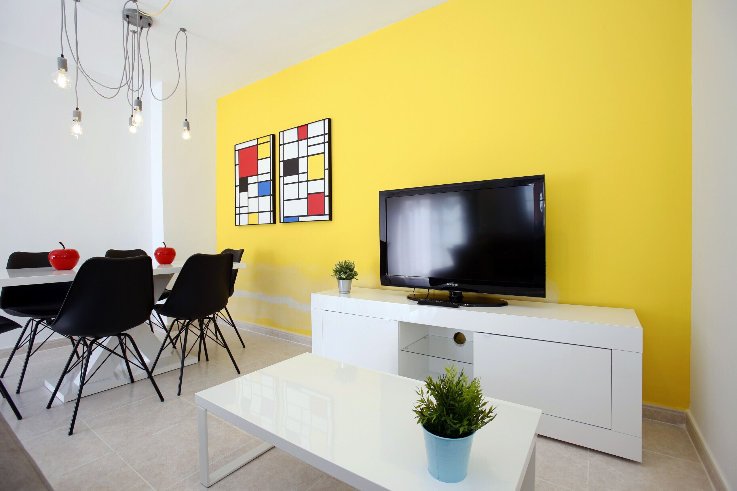 A modern living room with a yellow accent wall, a flat-screen TV, a white console, black chairs, and a glass coffee table with a plant.