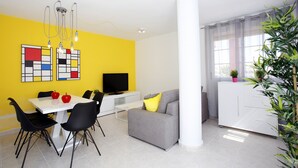 A modern living room with a yellow accent wall, a grey sofa, a white dining table with black chairs, and a white cabinet with a TV. - 177 Maravista (Denia)