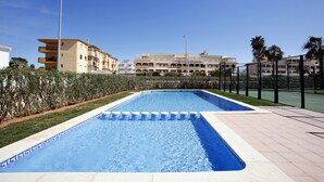 A rectangular swimming pool with a tiled edge, surrounded by a low hedge and a paved walkway. In the background, there are multi-story buildings with balconies and a clear blue sky. - 177 Maravista (Denia)