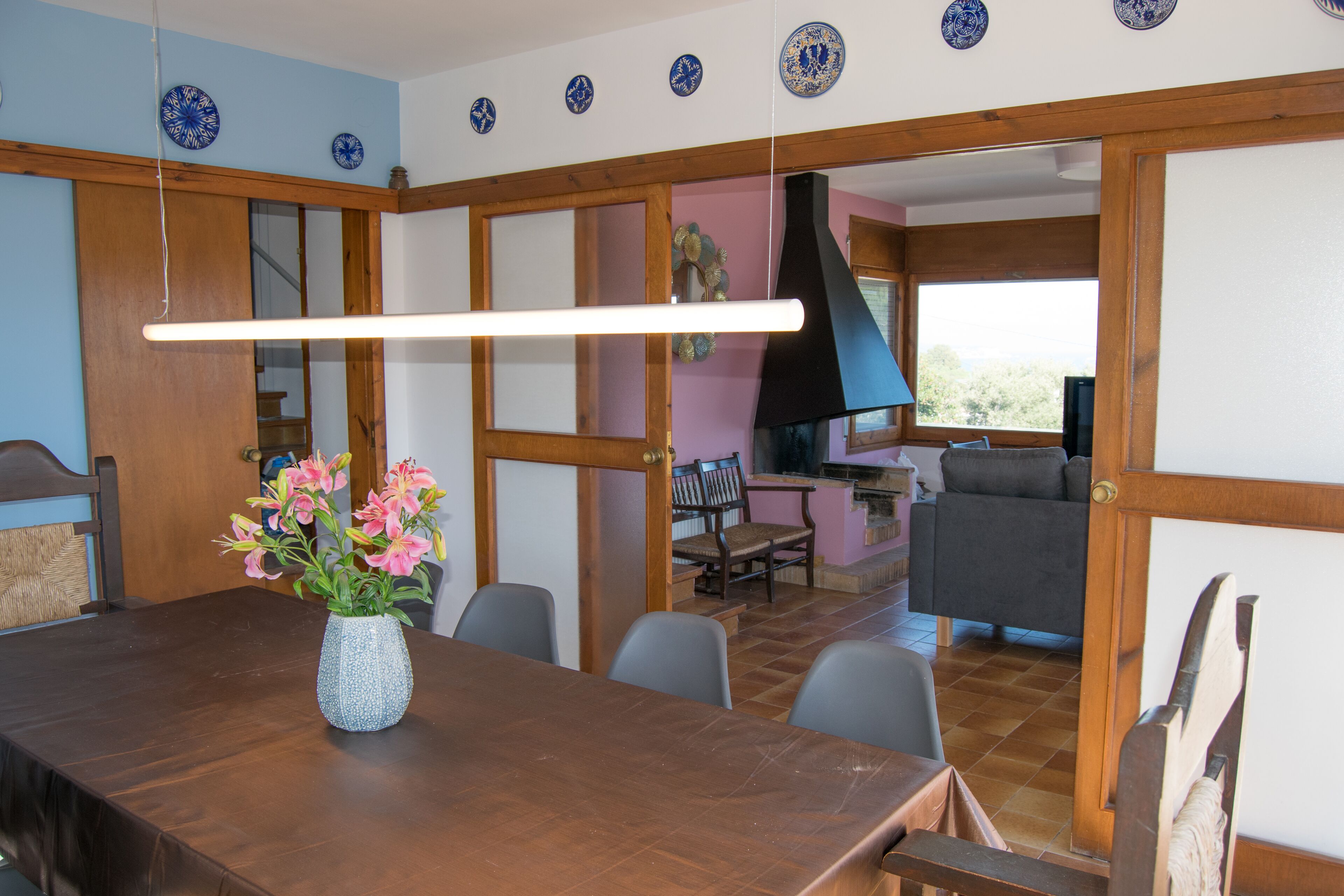 A dining area with a wooden table, chairs, and a vase of flowers. There is a kitchen area visible in the background with a black range hood and a window.