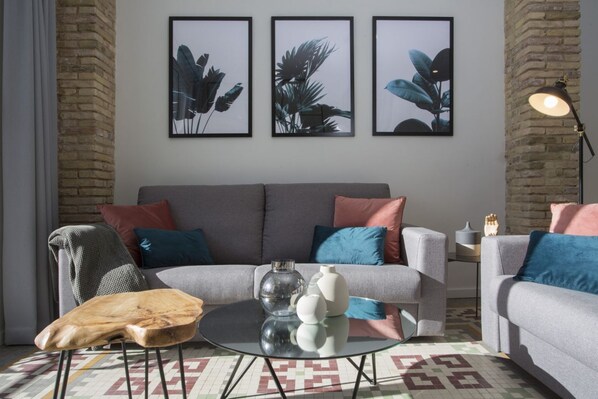 A modern living room with a grey sofa, colorful cushions, a round glass coffee table, and three framed botanical prints on the wall.