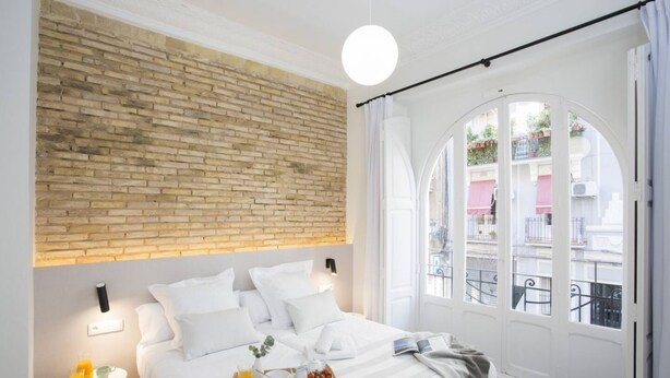 A hotel room with a brick wall, a bed with white bedding, a breakfast tray, and a view of the outside balcony. - Singular Stays Parque Central IX (Valencia)