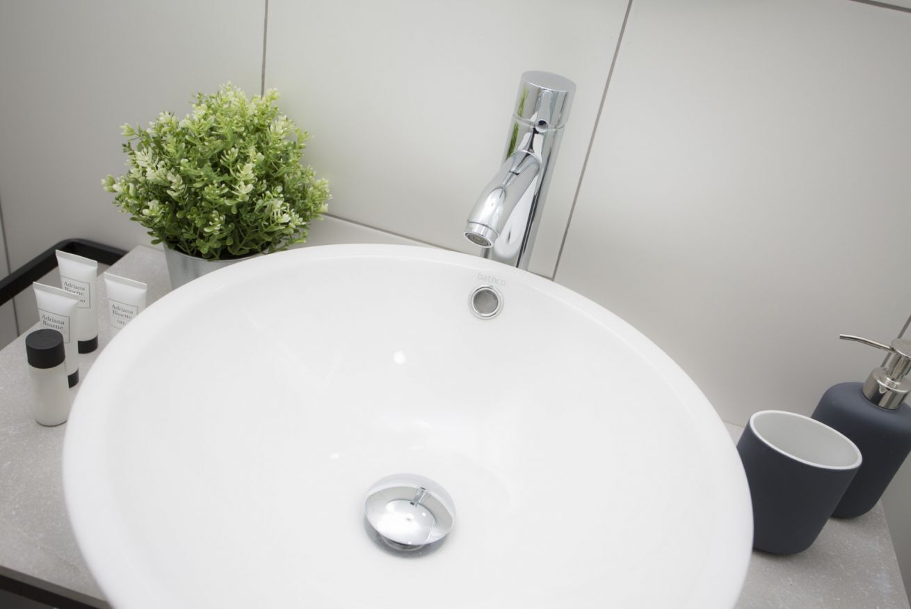 Un baño con lavamanos blanco, grifo cromado, una pequeña planta en maceta y artículos de aseo en la encimera.