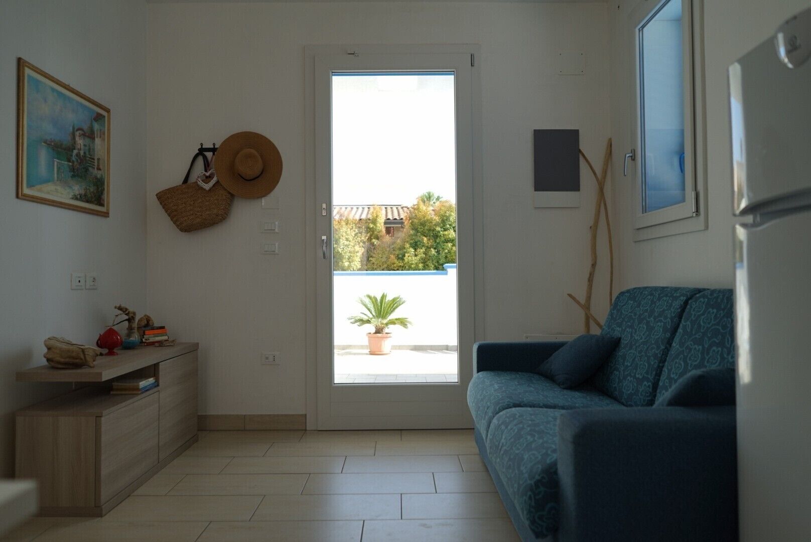 Ein Wohnzimmer mit einem blauen Sofa, einem Holzschrank, einem Hut an der Wand und einer Tür, die auf einen Balkon mit einem Blumentopf führt.
