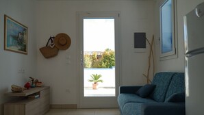 Una sala de estar con un sofá azul, un aparador de madera, un sombrero en la pared y una puerta que da a un balcón con una planta en maceta.