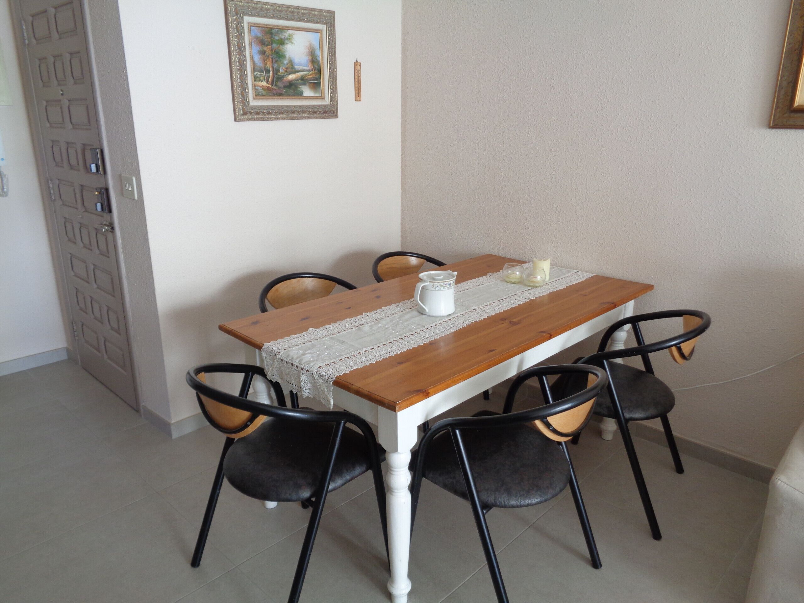 A dining table with a lace tablecloth, six chairs, and a painting on the wall.