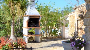 Un bâtiment en pierre avec un espace barbecue en briques, entouré de plantes en pot et d’un palmier.