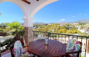 A balcony with a wooden table and chairs, a lantern, and a view of a town and mountains.