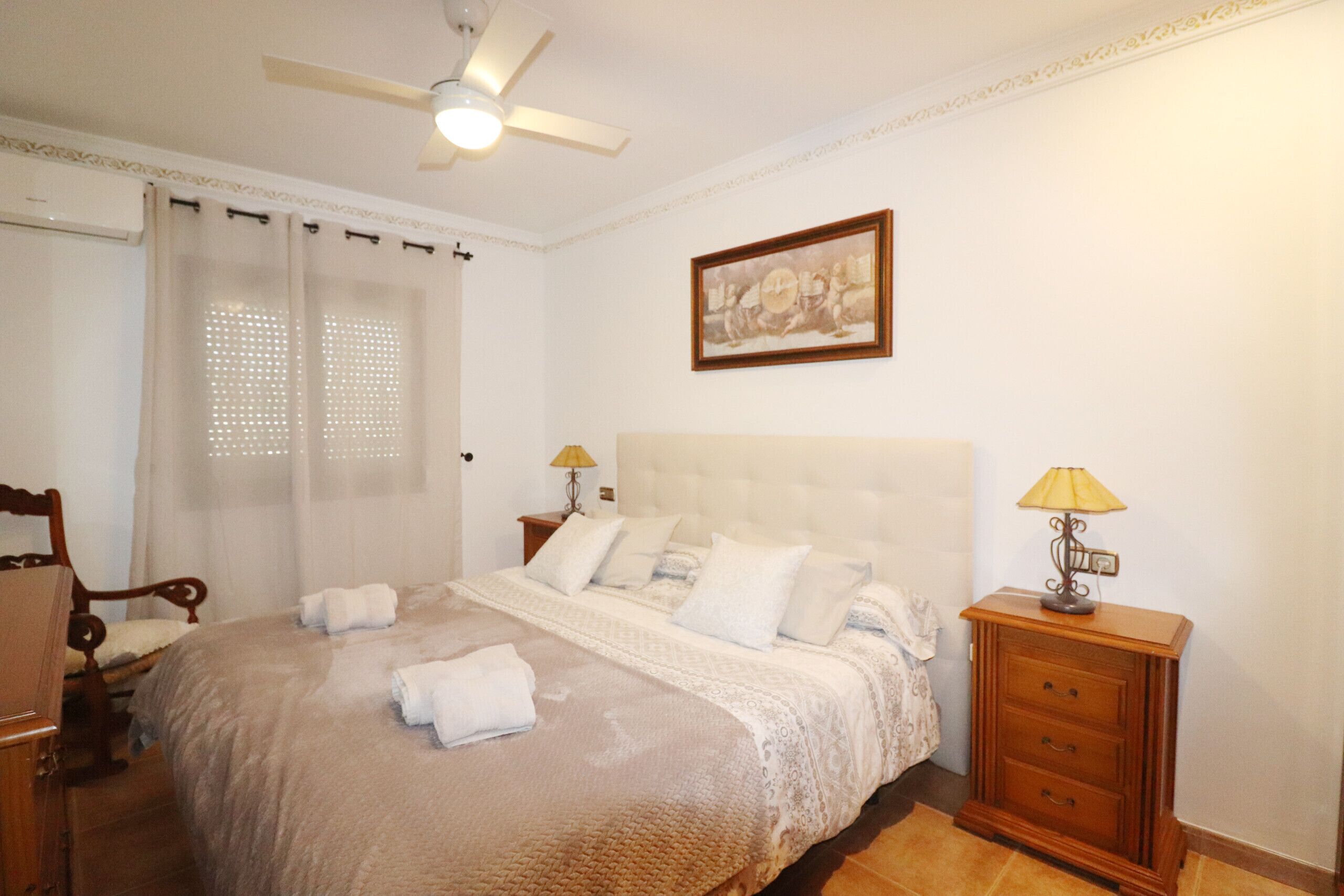 Ein Schlafzimmer mit Bett, Nachttisch, Stuhl, Deckenventilator und Wandlampe.