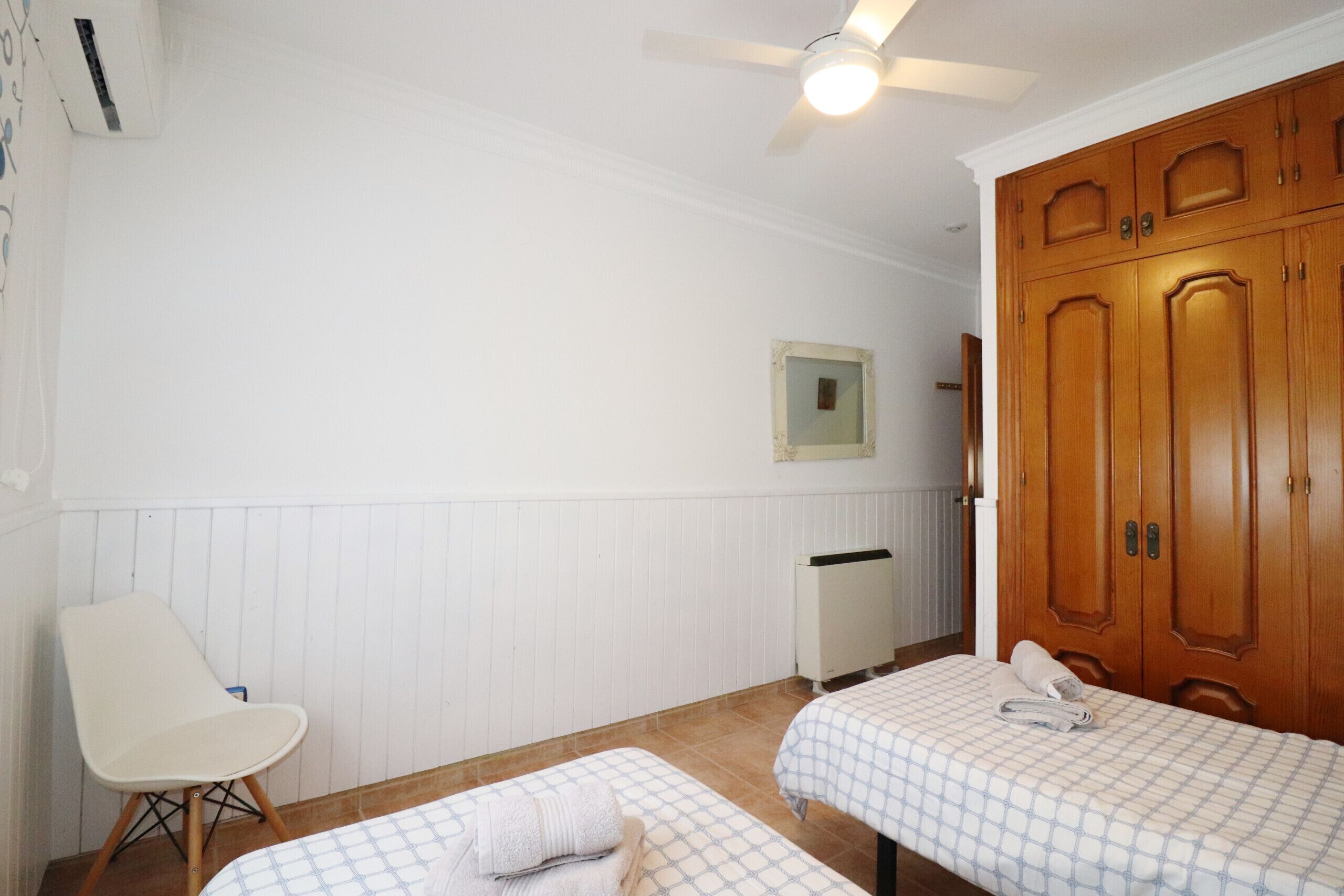 A room with two beds, a chair, a ceiling fan, wooden wardrobe, and a radiator.